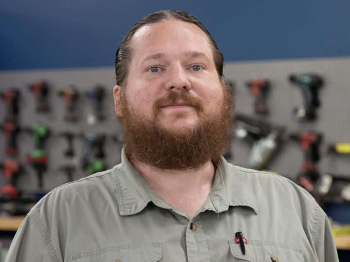 Man with a beard wearing a green shirt stands in front of a tool display on a blue wall.