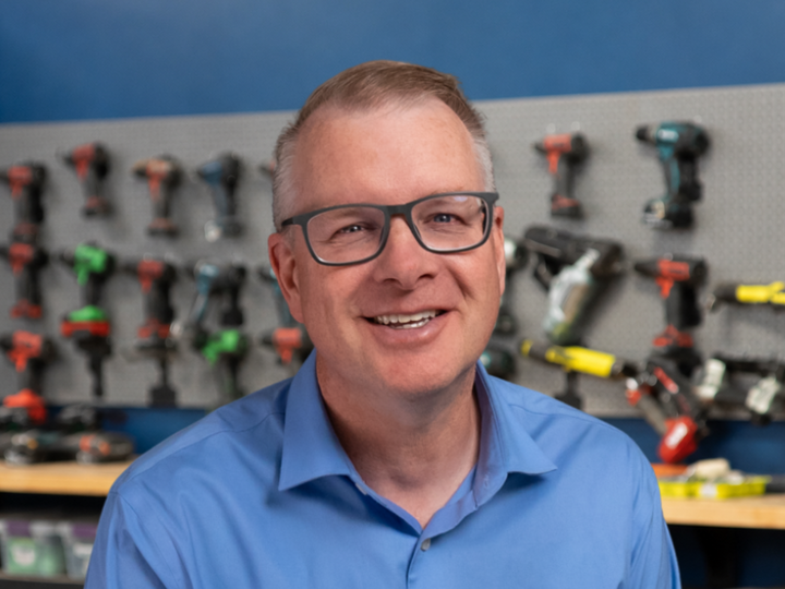 Man smiling, wearing glasses and a blue shirt, with a wall of tools hanging behind him.